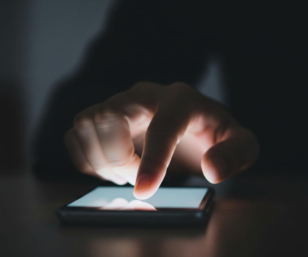 Dłoń z wyraźnie zarysowanym palcem wskazującym dotyka ekranu smartfona leżącego na płaskiej powierzchni. Ekran urządzenia świeci jasnym, niebieskawym światłem, które odbija się od opuszka palca i części dłoni, tworząc świetliste punkty i cienie. Tło jest ciemne i rozmyte, z niewyraźnymi konturami sugerującymi postać użytkownika.