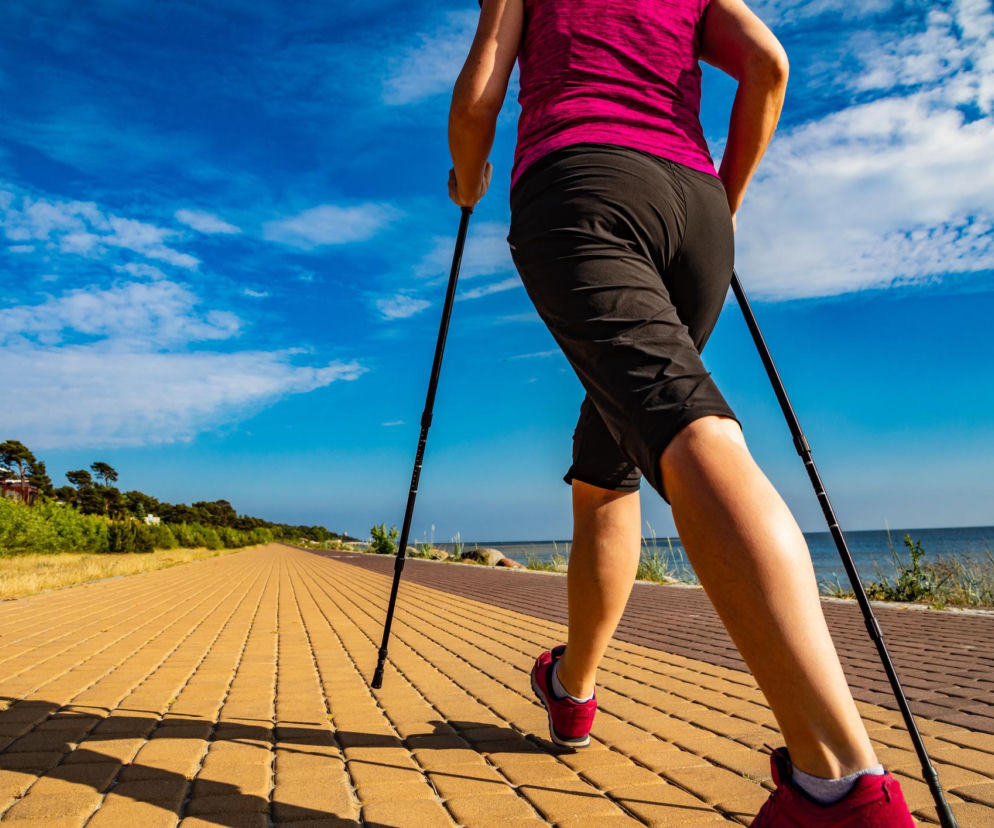 Osoba uprawiająca nordic walking maszeruje po nadmorskiej promenadzie w słoneczny dzień, z widokiem na morze i niebieskie niebo. To efektywny trening, o którym przeczytasz więcej na portalu Poradnik Zdrowie.