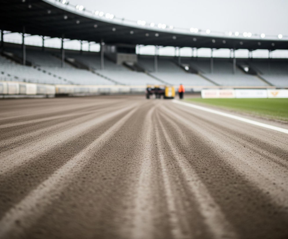 Piaskowa nawierzchnia toru z widocznymi, równoległymi śladami po grabieniu wypełnia większość kadru, rozciągając się od dołu do góry w oddali, gdzie zamazane kształty sugerują pojazd i postać. Po prawej stronie toru biegnie biała linia, oddzielająca go od pasa zielonej trawy, a za nią widoczne są niewyraźne, kolorowe bandy reklamowe. W tle wznosi się puste, betonowe trybuny stadionu pod częściowo zadaszoną konstrukcją, na której z daleka widać rzędy jasnych świateł.