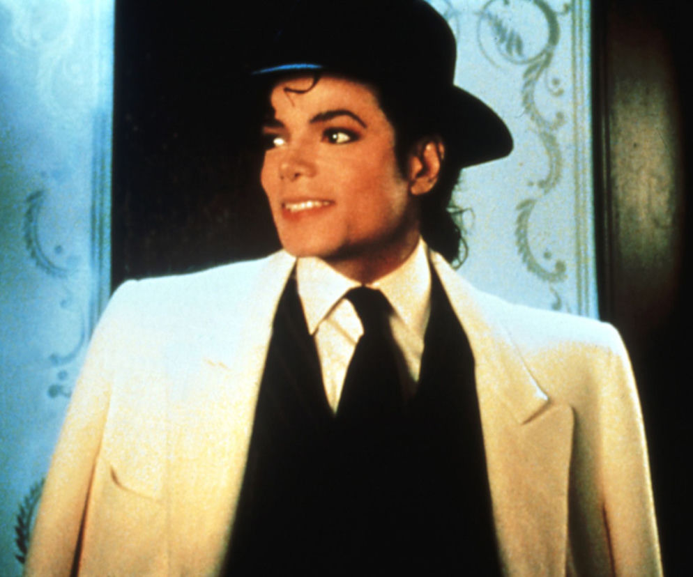 Michael Jackson w kapeluszu i białej marynarce uśmiecha się patrząc w bok. O procesie muzyka przeczytasz w serwisie Eska.