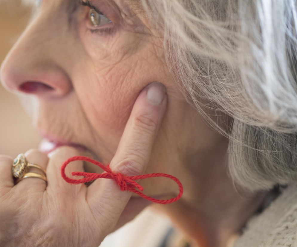 Profil starszej kobiety z czerwoną nitką zawiązaną na palcu. O wczesnej diagnostyce Alzheimera przeczytasz na Poradnik Zdrowie.