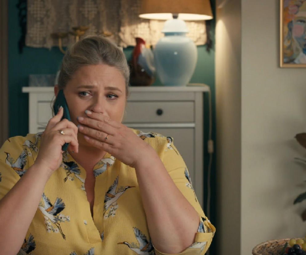 Aldona (Elżbieta Romanowska) z serialu Barwy szczęścia rozmawia przez telefon, zakrywając usta dłonią, co sugeruje szok i niedowierzanie. Tło to wnętrze pokoju z lampą i obrazem, tworzące domową atmosferę. Więcej o tym na Super Seriale.