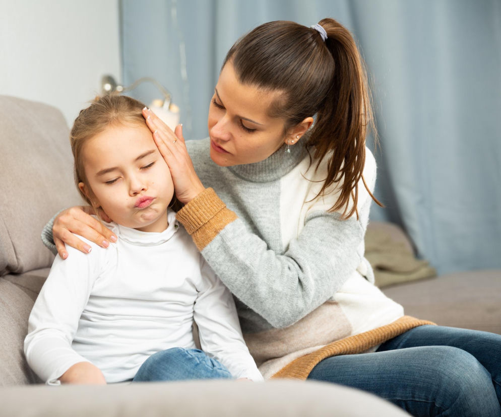 Zmartwiona kobieta delikatnie dotyka czoła i skroni dziewczynki, która z zamkniętymi oczami i wydętymi ustami sygnalizuje ból, siedząc obok matki na kanapie. Na Poradnik Zdrowie znajdziesz sposoby na łagodzenie bólu ucha u dziecka.