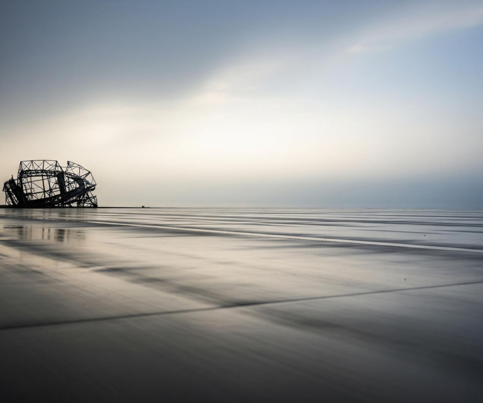 Na pierwszym planie, z lewej strony, widać ciemną, metalową konstrukcję o nieregularnym, kulistym kształcie, składającą się z wielu połączonych ze sobą prętów. Konstrukcja częściowo wystaje ponad linię horyzontu. Poniżej rozciąga się płaska, pozioma powierzchnia, przypominająca mokry lub odbijający światło bruk, z wyraźnymi poziomymi liniami i smugami, które tworzą efekt głębi i refleksów. Tło stanowi rozległe, zachmurzone niebo, które stopniowo jaśnieje od ciemnego błękitu po lewej stronie do jasnego, niemal białego odcienia po prawej. Całość utrzymana jest w chłodnej palecie barw, z dominacją szarości, błękitów i bieli.
