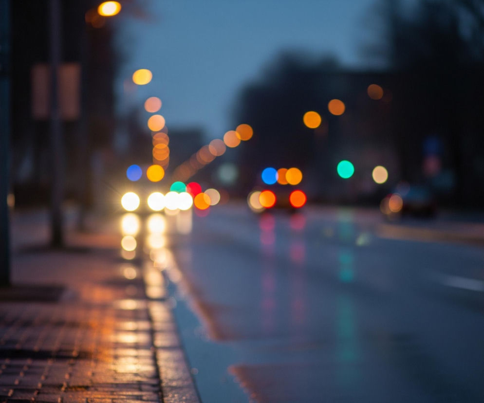 Uliczny widok przedstawia rozmyte światła miasta w deszczowy wieczór lub noc. Na pierwszym planie po lewej stronie znajduje się mokry chodnik z kostki brukowej, odbijający ciepłe, żółtopomarańczowe bliki od nieostrych źródeł światła. Po prawej stronie chodnika biegnie ciemna, mokra jezdnia, na której wyraźnie widać refleksy świateł pojazdów – jaskrawe, białe światła z przodu i czerwone z tyłu. W tle rozciąga się ciemne otoczenie, z kilkoma niewyraźnymi drzewami i budynkami, a nad nimi widać niebo w odcieniu granatu.