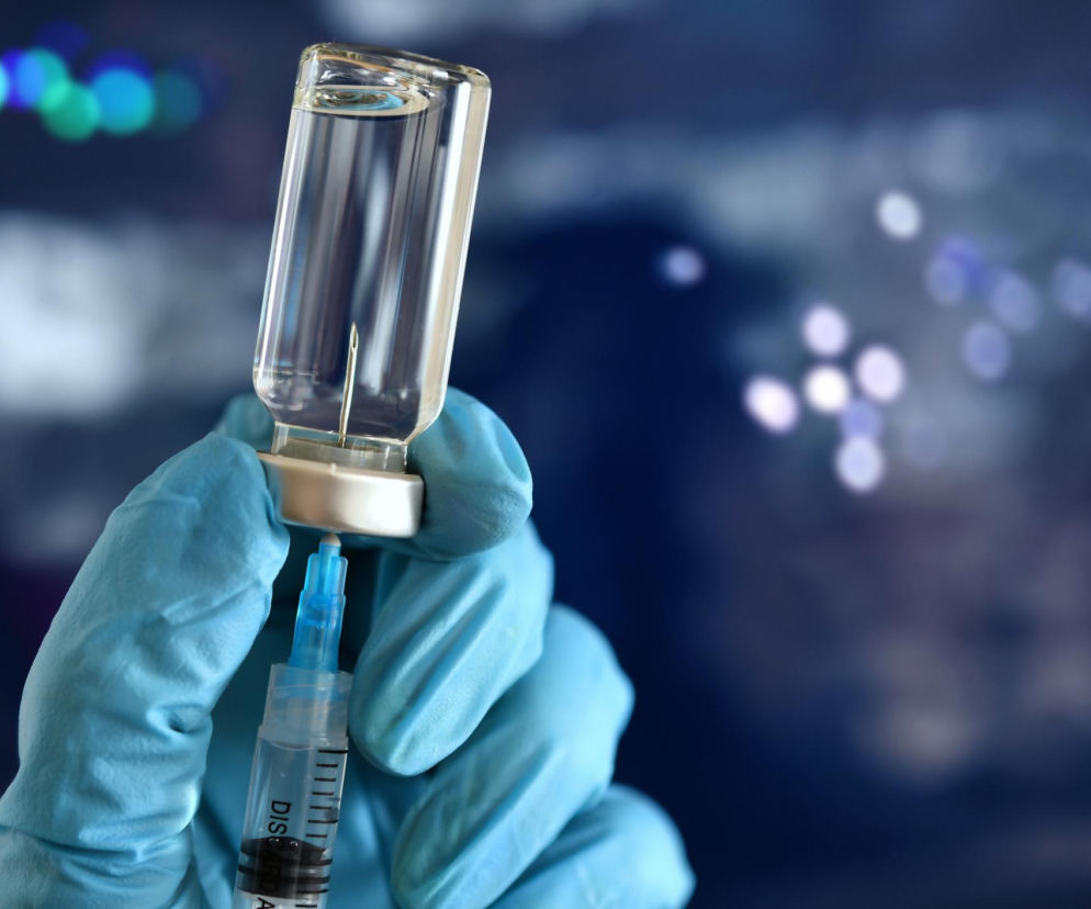 Dłoń w niebieskiej rękawiczce medycznej trzyma ampułkę ze szczepionką, z której wyciągana jest substancja do strzykawki. Scena symbolizuje kluczowe znaczenie szczepień w historii medycyny, o czym możesz przeczytać na portalu Poradnik Zdrowie.