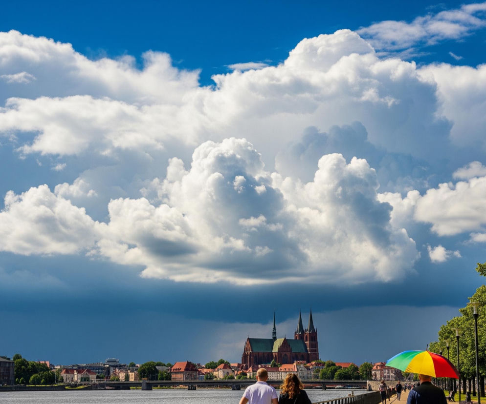 Nad rzeką rozciąga się panorama miasta z charakterystycznym, dużym kościołem o dwóch spiczastych wieżach i ciemnoczerwonych murach, widocznym w centralnej części zdjęcia. Niebo jest częściowo błękitne, a częściowo pokryte ogromnymi, białymi chmurami kłębiastymi, które w dolnych partiach przybierają ciemniejsze, burzowe odcienie. Na pierwszym planie, po prawej stronie, widać aleję obsadzoną rzędem drzew o jasnozielonych liściach, a wzdłuż niej spacerują ludzie; jedna osoba wyróżnia się trzymając wielobarwny parasol w kolorach czerwonym, żółtym, zielonym i niebieskim.