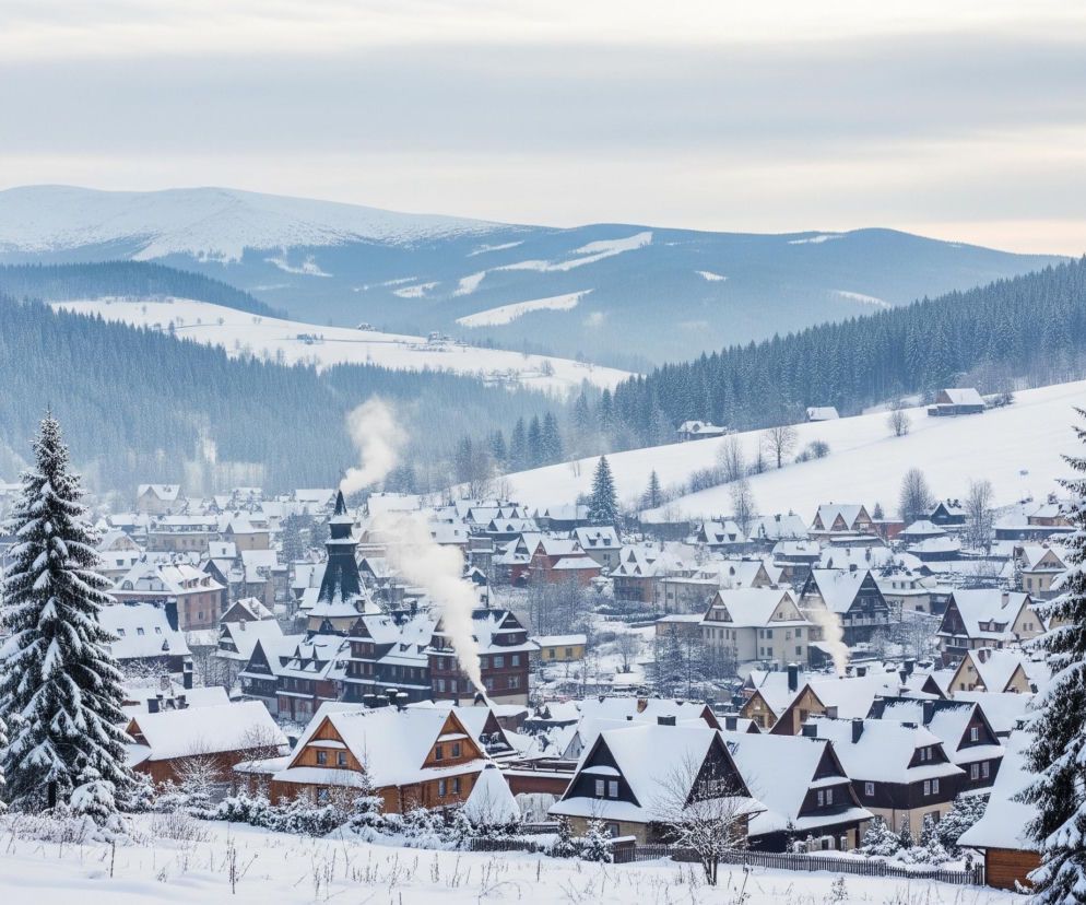 Malowniczy, zimowy krajobraz przedstawia zaśnieżone miasteczko położone w dolinie, otoczone gęstymi lasami iglastymi i wysokimi, pokrytymi śniegiem górami. Dwa okazałe, ośnieżone drzewa iglaste dominują na pierwszym planie, jedno po lewej, drugie po prawej stronie, kadrując widok na osadę. Domy w miasteczku mają dachy pokryte grubą warstwą białego śniegu, a z kominów unosi się biały dym, świadczący o paleniu w piecach. W tle rozciągają się kolejne rzędy zalesionych wzgórz, a nad nimi widać masywne, białe szczyty górskie pod jasnym, zachmurzonym niebem.