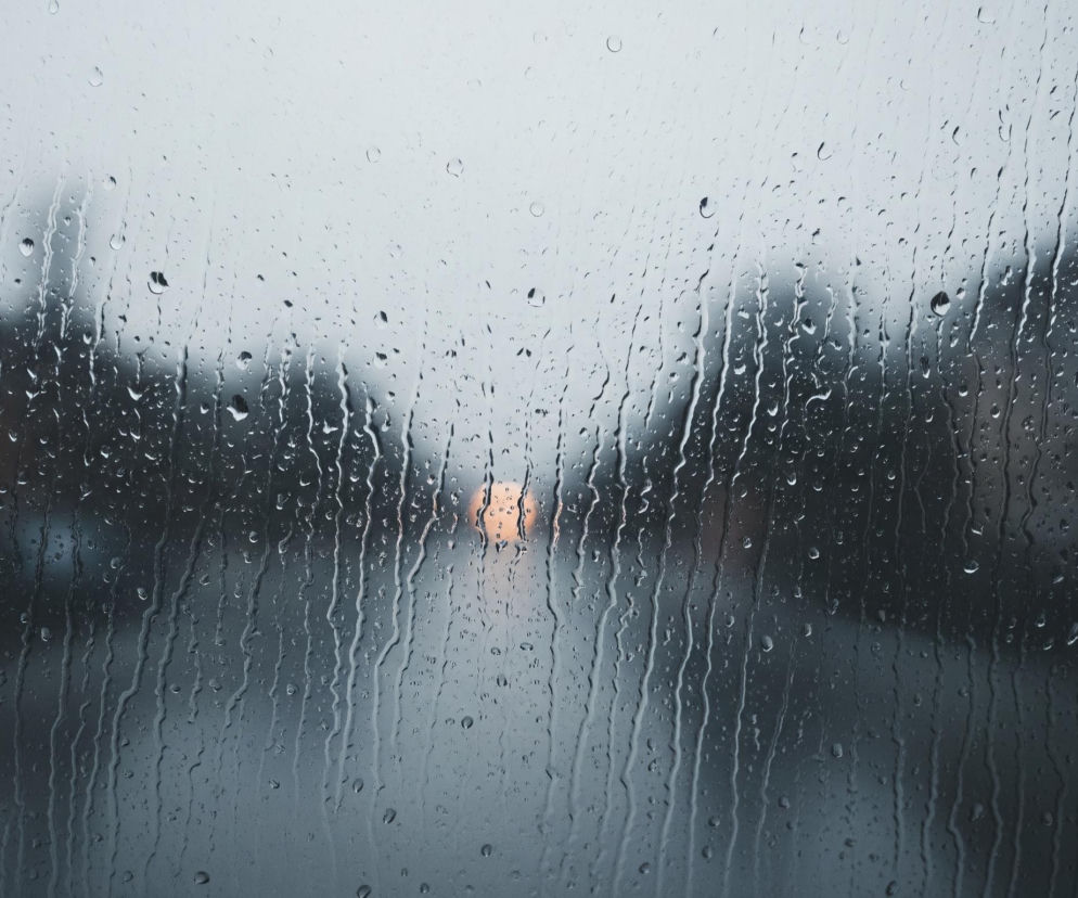 Szyba pokryta licznymi kroplami deszczu zajmuje całą powierzchnię obrazu, tworząc rozmyty widok tła. Na szybie widać pionowe smugi wody spływającej w dół oraz pojedyncze, okrągłe krople. W tle, rozmazane sylwetki drzew lub budynków o ciemnych, szaro-zielonych barwach, wyłaniają się zza warstwy wody. Centralnie, w oddali, dostrzegalne jest jasne, pomarańczowo-żółte światło, prawdopodobnie reflektor pojazdu.