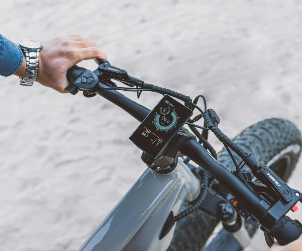 Mężczyzna w koszuli jeansowej ze srebrnym zegarkiem na nadgarstku prowadzi rower elektryczny. Widoczne jest jego ujęcie kierownicy z wyświetlaczem. Jazda na rowerze może zafałszować wynik badania PSA, o czym przeczytasz na Poradnik Zdrowie.