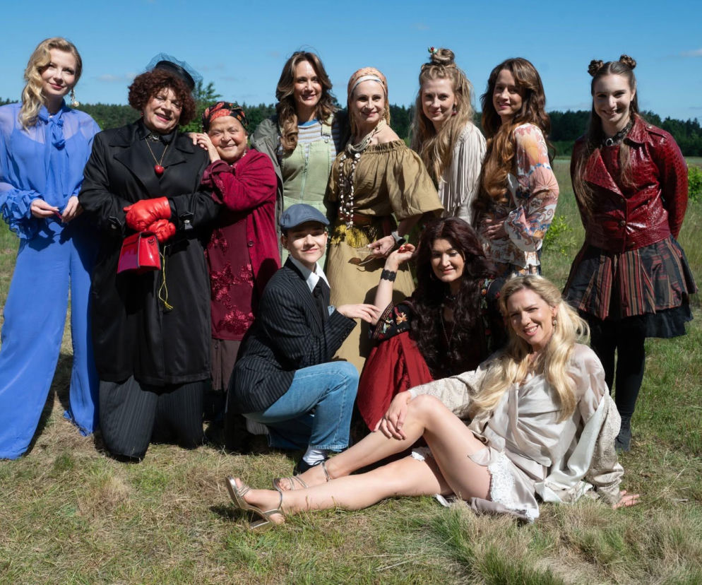 Grupa aktorek z serialu Ranczo, w tym znane bliźniaczki, pozuje na tle otwartego terenu z drzewami. Kobiety w różnorodnych strojach filmowych, w tym jedna w niebieskim kombinezonie, inna w czarnym płaszczu z czerwonymi rękawiczkami. O nowych twarzach w serialu przeczytasz na naszym portalu.