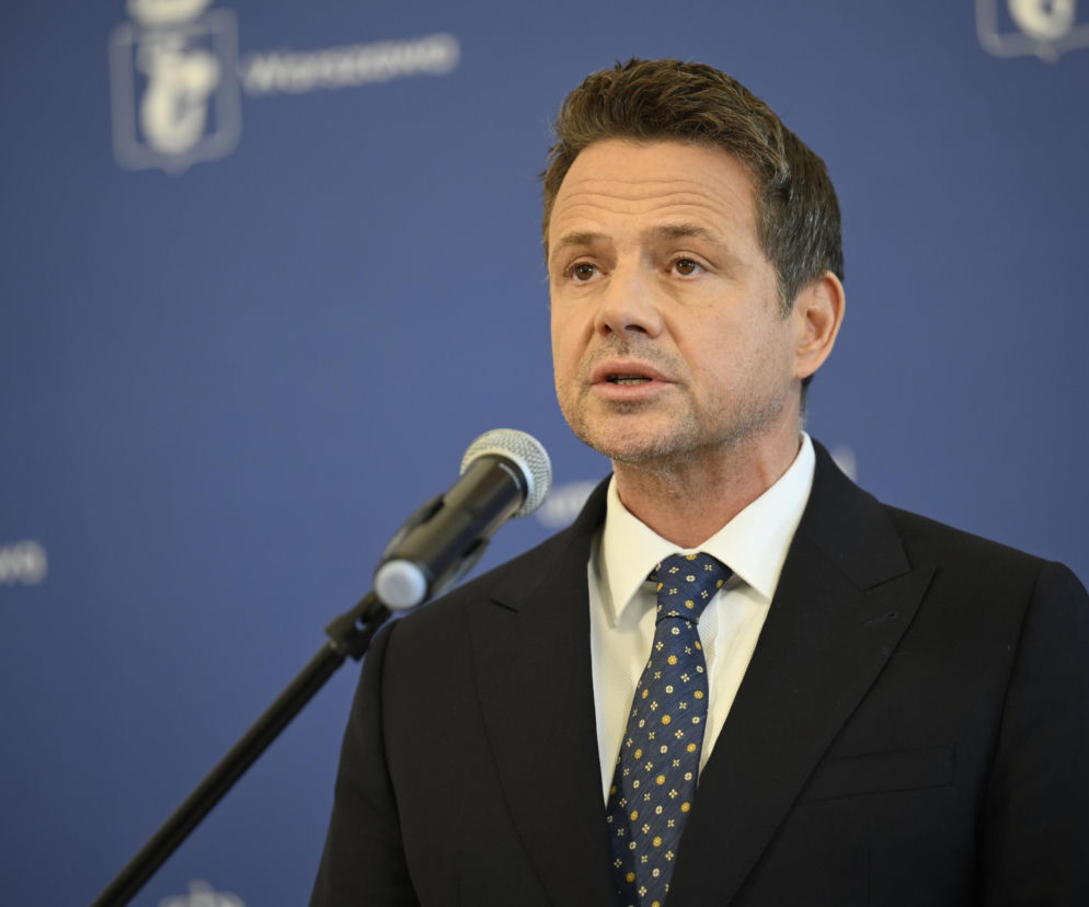 Rafał Trzaskowski przemawiający do mikrofonu na tle niebieskiej ściany z białymi, niewyraźnymi logotypami. Polityk ubrany jest w garnitur, białą koszulę i krawat w niebiesko-żółty wzór. Temat porażki Trzaskowskiego w wyborach prezydenckich przeczytasz na naszym portalu.
