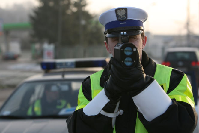 Funkcjonariusz polskiej drogówki w mundurze z tarczą na czapce i odblaskową kamizelką, mierzący prędkość pojazdów policyjnym radarem. Scena nawiązuje do ogólnopolskiej akcji „Alkohol i narkotyki”. Więcej o kontrolach na drogach przeczytasz na Super Biznes.