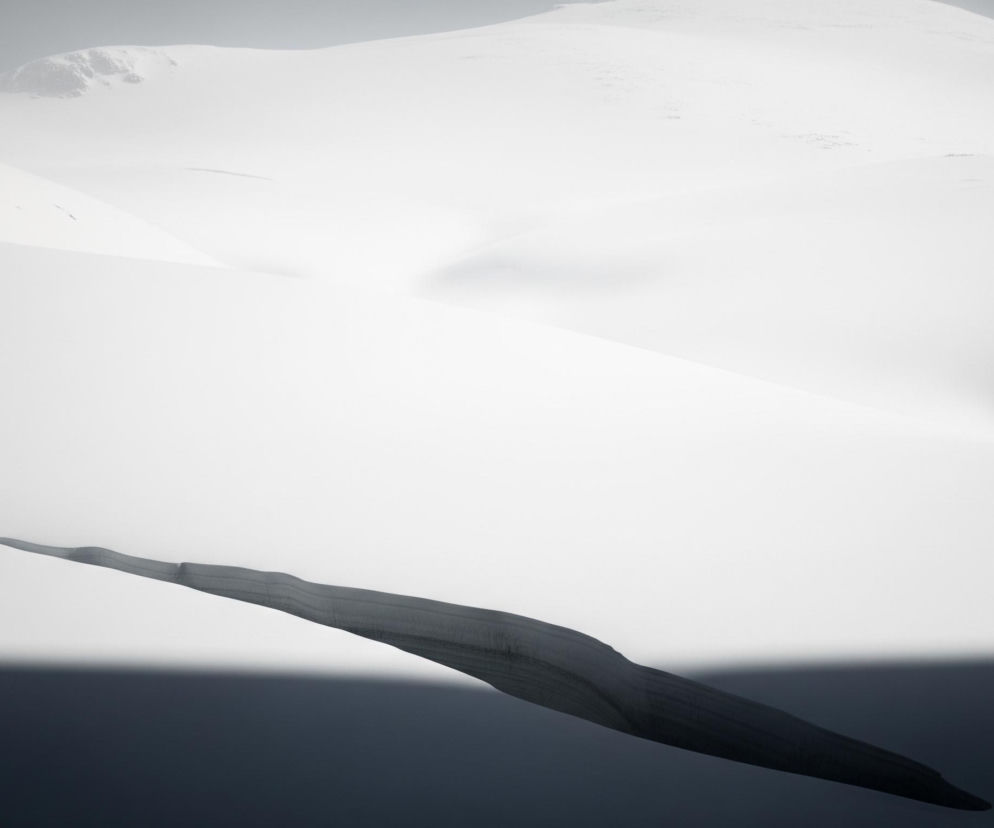 Zasypany śniegiem, szeroki krajobraz górski dominuje w kadrze, z wyraźnym pęknięciem lub szczeliną na pierwszym planie. Obszar ten jest pokryty jednolitą, białą warstwą śniegu, przeplataną subtelnymi cieniami, które nadają rzeźbie terenu głębię i zróżnicowanie. Szarobłękitny, ciemny obszar znajduje się w dolnej części obrazu, częściowo zakryty przez wydłużony, ciemny cień, który ciągnie się od lewej dolnej krawędzi w kierunku środka, tworząc dynamiczną linię. Na horyzoncie widoczne są zamglone, rozległe szczyty górskie, co sugeruje dużą przestrzeń i odległość, a ich kształty zlewają się z jasnym, prawie białym niebem.