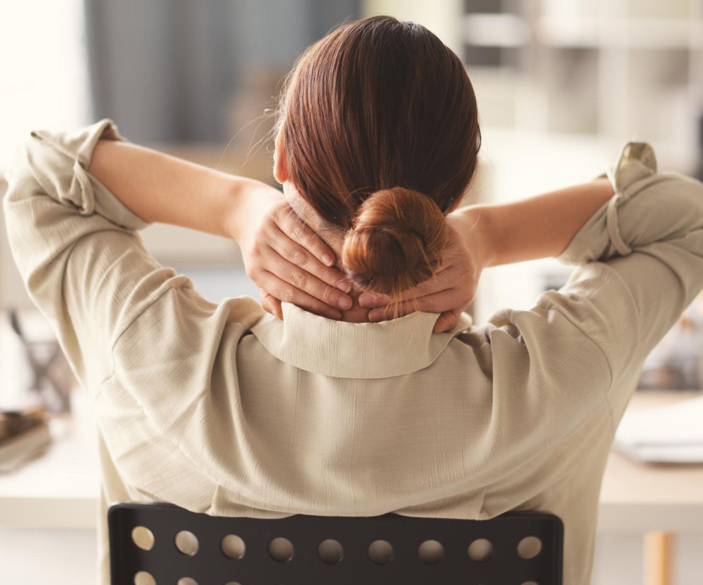 Kobieta z ciasno upiętym kokiem, w beżowej koszuli, siedząca tyłem do obiektywu, masująca swój kark obiema rękami. Taki rodzaj bólu karku, często wynikający ze stresu, dotyka coraz więcej Polaków, o czym przeczytasz na Poradnik Zdrowie.