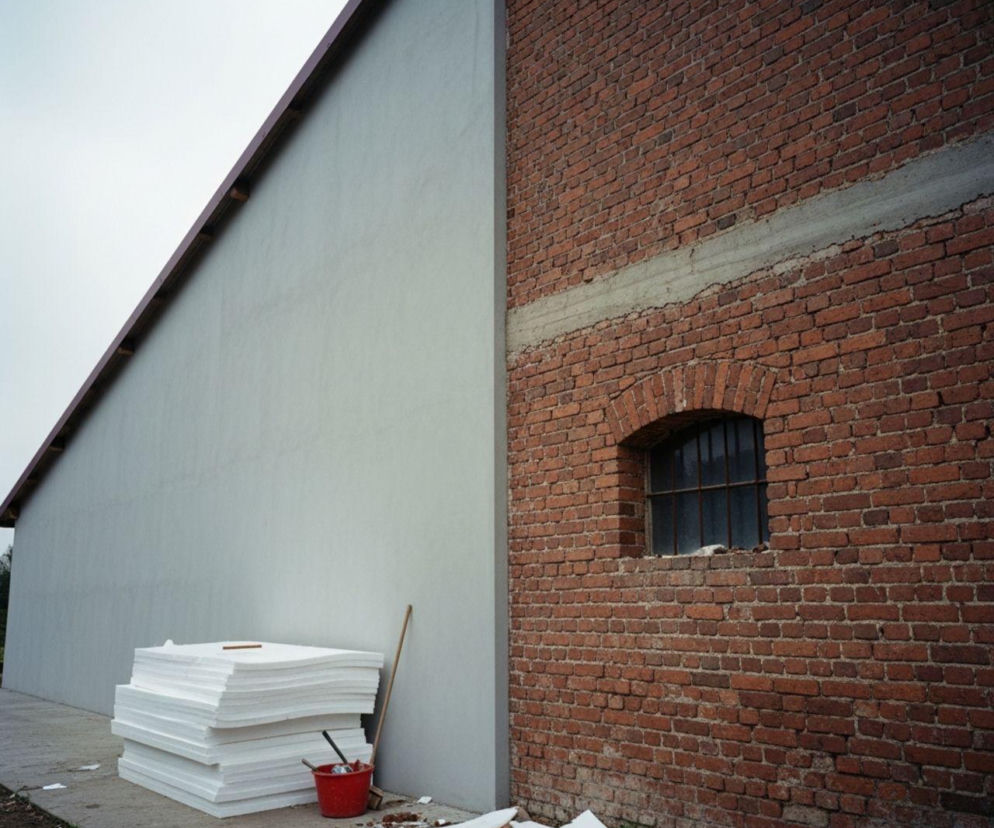 Termomodernizacja budynku gospodarczego. Białe płyty styropianowe i czerwone wiadro stoją przy ścianie z jasnego tynku, obok ceglanej fasady z łukowatym oknem. Temat dotacji na ocieplenie budynków rolniczych na Super Biznes.