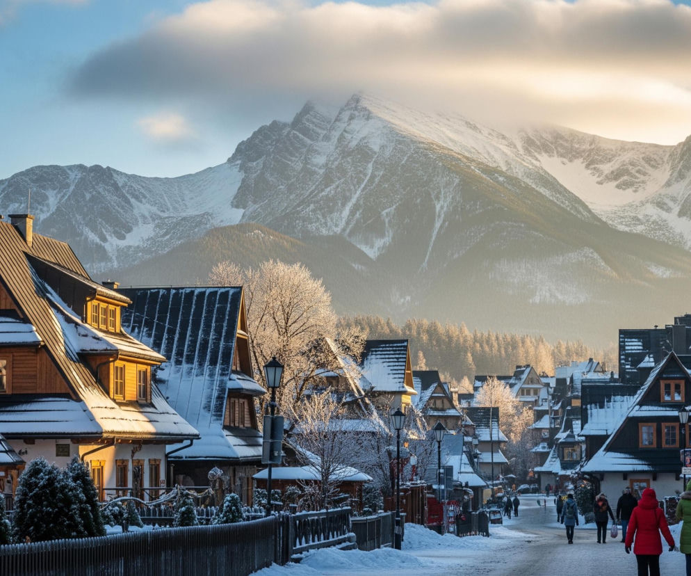 Zimowy krajobraz przedstawia zaśnieżoną ulicę w górskiej miejscowości, otoczoną domami o drewnianych fasadach i spadzistych dachach pokrytych białym puchem. W tle dominują majestatyczne, ośnieżone szczyty górskie, częściowo zasłonięte przez kłębiaste, jasne chmury, oświetlone promieniami słońca. Po lewej stronie widoczne są gęste drzewa iglaste, również pokryte śniegiem i szronem, a wzdłuż ulicy stoją latarnie uliczne i poruszają się nieliczni piesi.