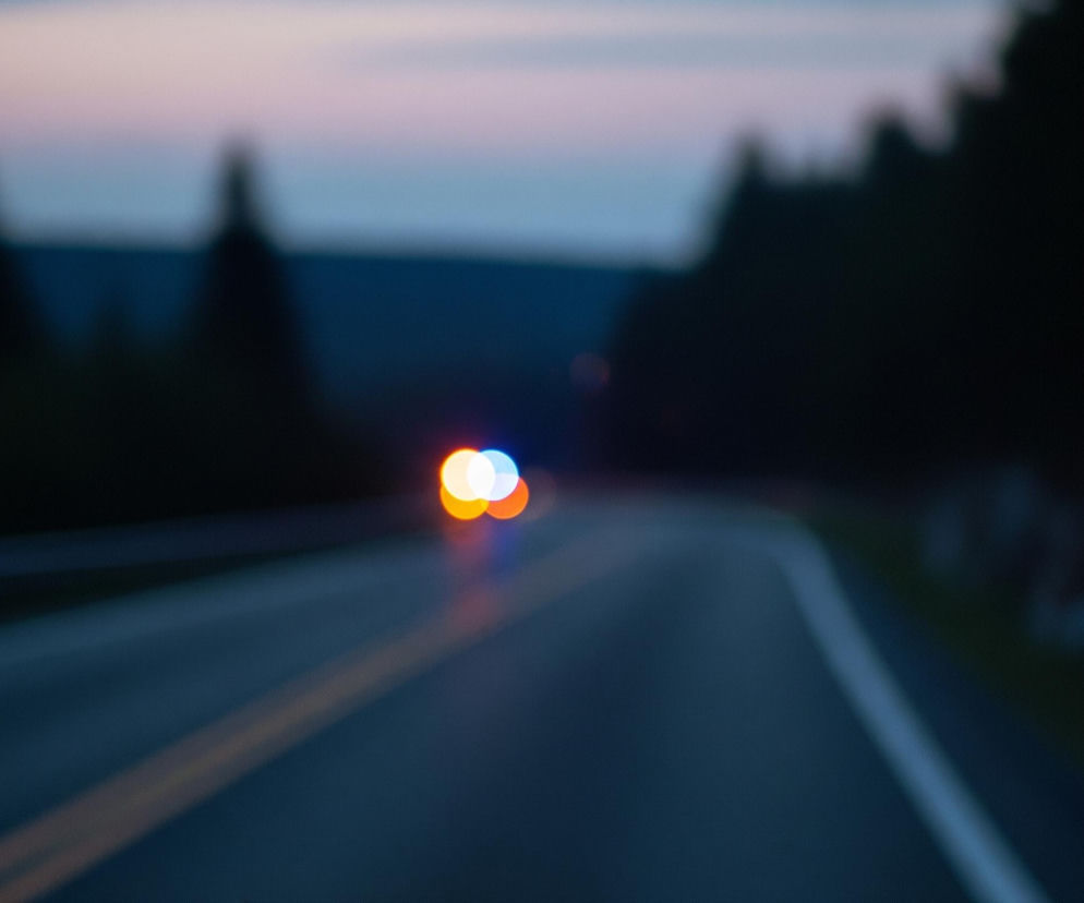 Rozmazane zdjęcie przedstawia drogę asfaltową w zmierzchu lub w nocy, z niewyraźnymi światłami pojazdu w oddali. Na pierwszym planie widoczna jest ciemna, gładka powierzchnia jezdni, z wyraźną, jasną, przerywaną linią po prawej stronie oraz ciągłą, podwójną, żółtą linią bliżej środka kadru. W tle, na horyzoncie, rozpościera się ciemny pas lasu, nad którym widoczne jest niebo w odcieniach fioletu, różu i błękitu, sugerujące zachód słońca lub świt. Pośrodku obrazu, w oddali, znajdują się rozmyte kule światła w kolorach białym, pomarańczowym i niebieskim, przypominające reflektory i lampy sygnalizacyjne samochodu.