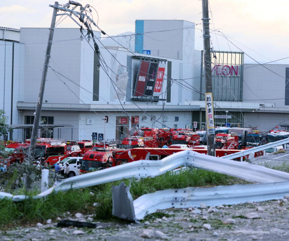 Zniszczona barierka i słup energetyczny w Japonii. W tle wozy strażackie. O skutkach trzęsienia ziemi przeczytasz w Eska.