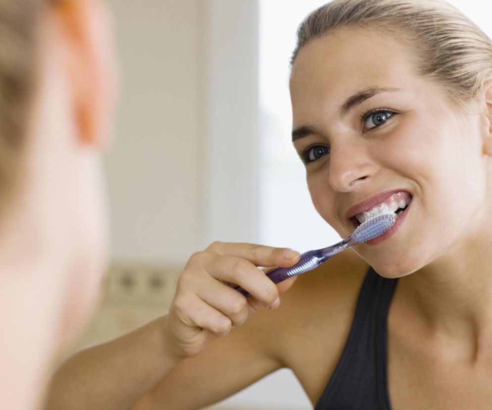 Uśmiechnięta młoda kobieta z blond włosami spiętymi w kok, patrząca w lustro i myjąca zęby szczoteczką z widoczną pastą. Twarz odbija się w lustrze, wskazując na codzienną higienę jamy ustnej. Więcej o prawidłowym myciu zębów przeczytasz na Poradnik Zdrowie.