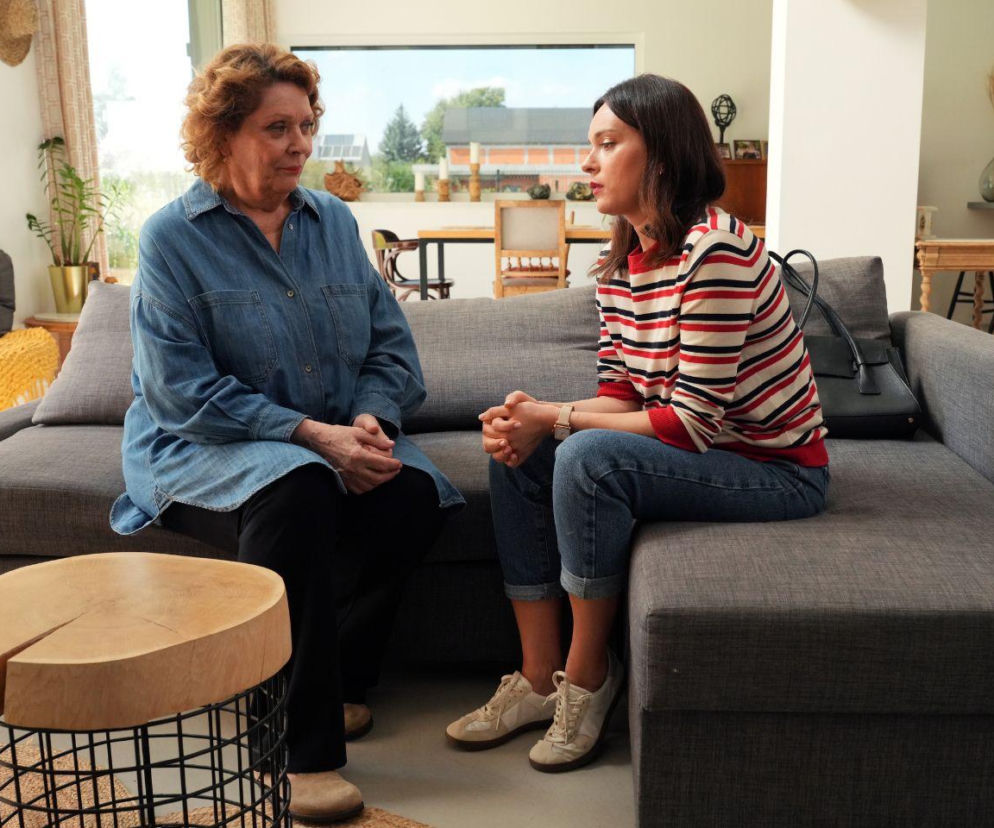 Dwie kobiety, starsza w dżinsowej koszuli i młodsza w pasiastym swetrze, siedzą na szarej kanapie w jasnym salonie, prowadząc rozmowę. Widok z serialu Barwy szczęścia dostępny na Super Seriale.