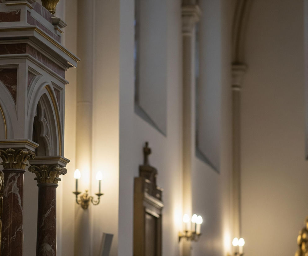 Złota figurka brodatego mężczyzny w długiej szacie z książką i krzyżem w jednej dłoni oraz uniesioną dłonią w geście błogosławieństwa, stoi w niszy ozdobionego kolumnami o marmurowym wzorze i złotymi detalami o gotyckich łukach, umieszczona z lewej strony kadru. W tle widać kolumny i ściany o jasnym, kremowym odcieniu, na których wiszą zapalone, dwuramienne kinkiety. Po prawej stronie, w głębi, majaczy rozmazany fragment kolejnego złotego obiektu, a wysoko, w prawym górnym rogu, widoczny jest duży, kolorowy witraż z postaciami świętych, oświetlony naturalnym światłem z zewnątrz.
