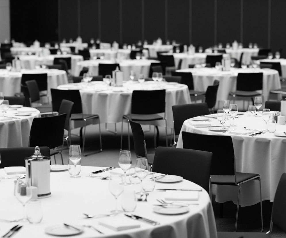 Wielka sala bankietowa jest wypełniona okrągłymi stołami, nakrytymi białymi obrusami, każdy z nich jest przygotowany do serwowania posiłków. Na stołach widać sztućce, białe talerze oraz różnego rodzaju szklanki i kieliszki do napojów. Przy każdym stole ustawione są czarne krzesła z metalowymi, połyskującymi nóżkami, czekające na gości. Całość utrzymana jest w czarno-białej kolorystyce, co nadaje scenie eleganckiego i formalnego charakteru.