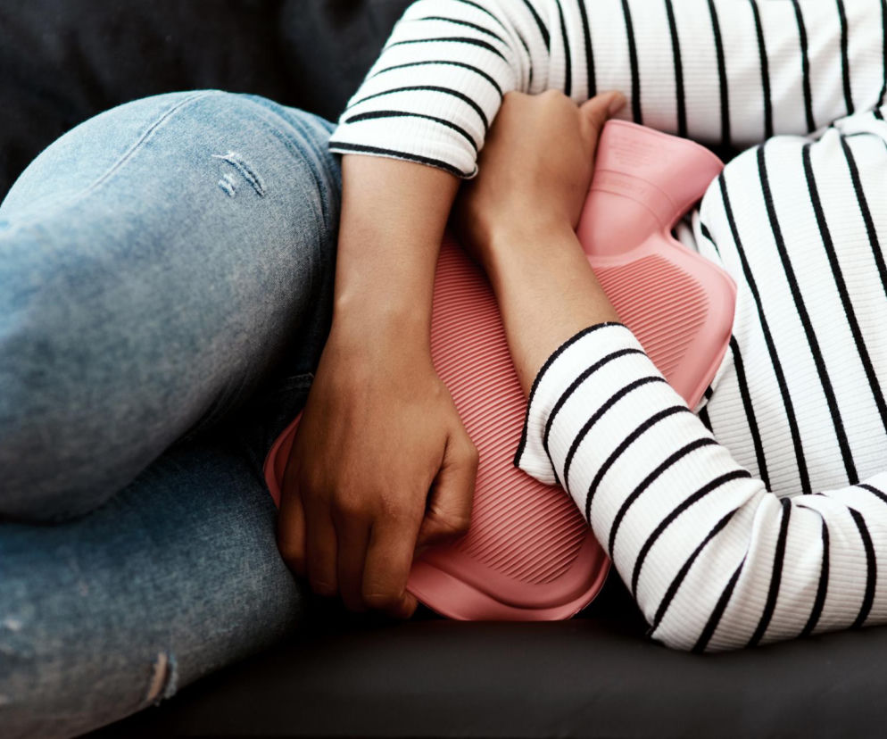 Kobieta leżąca na kanapie z termoforem na brzuchu, trzymająca ręce na rozgrzewającym okładzie, co symbolizuje łagodzenie bólu menstruacyjnego lub bólu towarzyszącego endometriozie. Na Poradnik Zdrowie można przeczytać o nowym teście krwi na endometriozę.