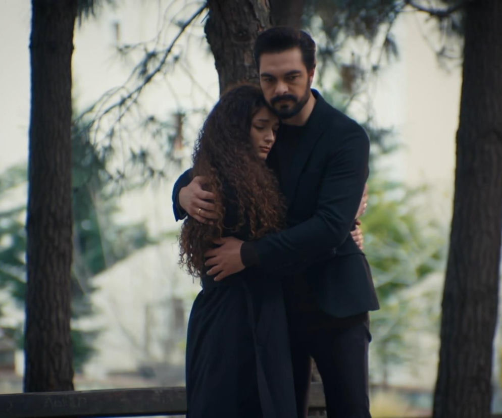 Nana i Yaman obejmują się, stojąc pod drzewami. Kobieta ma długie, kręcone włosy i jest ubrana na czarno, zamyka oczy, przytulając się do mężczyzny. Yaman w czarnym garniturze patrzy przed siebie z zatroskaną miną. Zdjęcie ilustruje odcinek serialu Dziedzictwo, o którym przeczytasz więcej na Super Seriale.