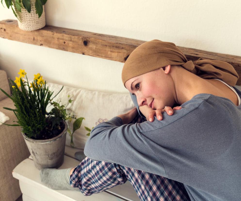 Młoda kobieta w beżowej chuście na głowie, symbolizującej leczenie onkologiczne, siedzi w zamyśleniu, obejmując kolana. Scena ukazuje pacjentkę zmagającą się ze szpiczakiem plazmocytowym, o czym można przeczytać na portalu Poradnik Zdrowie. Tło stanowią łóżko i drewniana półka z roślinami.