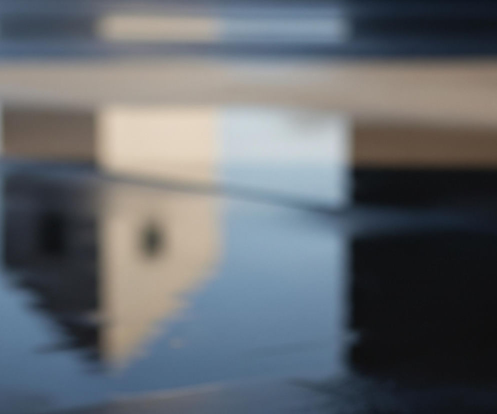 Rozmazany obraz przedstawia odbicie jasnej, prostokątnej formy z ciemnym otworem w górnej części, prawdopodobnie wieży lub wysokiego budynku, w lustrzanej, ciemnoniebieskiej powierzchni, która zajmuje dolną połowę kadru. Powyżej odbicia, w tle, widoczne są poziome pasy o zmiennej jasności: jeden jasny, beżowy pas w centrum, nad którym rozciąga się ciemniejszy, szarobrązowy obszar. Całość tworzy abstrakcyjną kompozycję barwnych smug i rozmytych kształtów, z dominantą błękitów, beżów i ciemnych brązów.