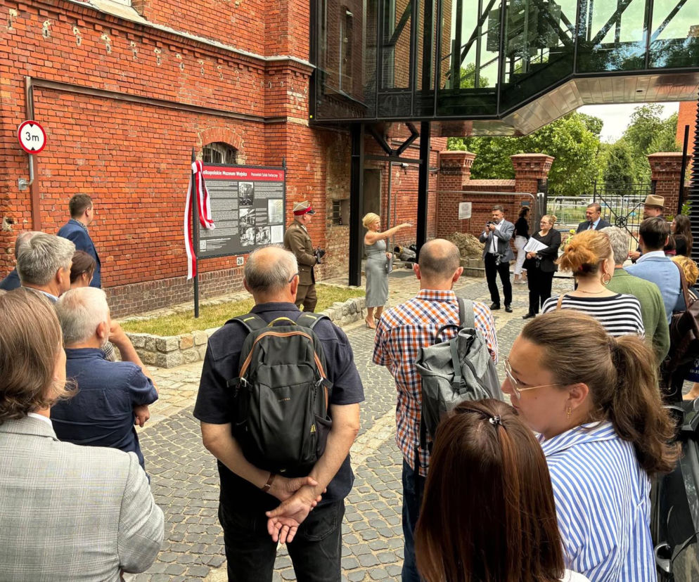 Grupa ludzi przy zabytkowym murze z czerwoną cegłą. O nowej tablicy Szlaku Fortecznego przeczytasz na Eska Poznań.