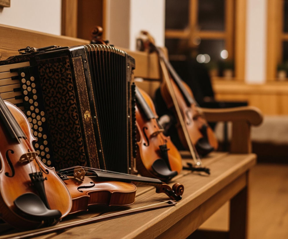 Na drewnianej ławce, w centralnej części zdjęcia, leży kilka instrumentów muzycznych. Na pierwszym planie, z lewej strony, widoczne są skrzypce o brązowo-pomarańczowym korpusie i czarnym podbródku, częściowo zasłaniające mniejsze skrzypce leżące obok. Obok nich znajduje się czarny akordeon z dekoracyjnymi, złotymi wzorami i białymi klawiszami oraz guzikami. Dalej w tle, na ławce, ustawione są kolejne skrzypce oraz większy instrument smyczkowy, prawdopodobnie wiolonczela, wszystkie o podobnej, brązowej kolorystyce. Tło stanowią drewniane panele ścian i okno z widocznymi rozmazanymi światłami w oddali, a także czarny statyw do nut po prawej stronie.