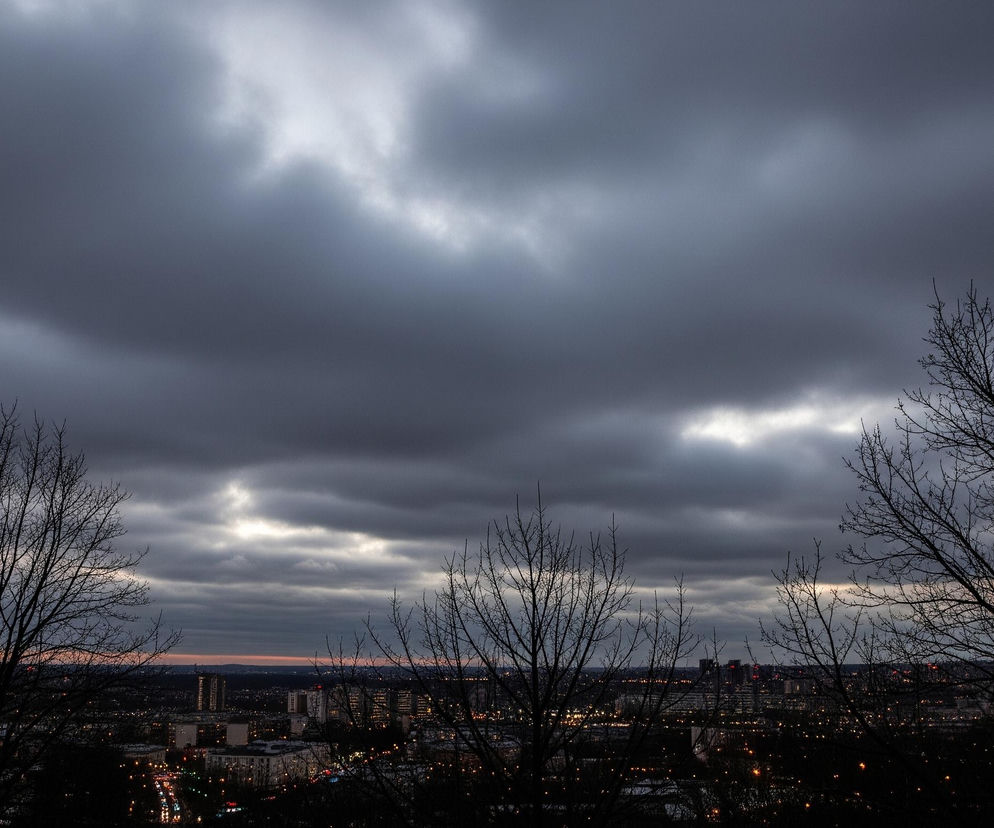 Ciemne, szare chmury dominują niebo, z jaśniejszymi smugami światła przebijającymi się przez nie w kilku miejscach, głównie w centrum i po prawej stronie. Na horyzoncie, pod chmurami, rozciąga się miejski pejzaż z licznymi, rozświetlonymi budynkami i ulicami, widoczne są światła samochodów. Przed miastem, na pierwszym planie, znajdują się sylwetki nagich drzew z licznymi gałęziami, częściowo zasłaniające widok na zabudowania. W lewej dolnej części obrazu można dostrzec rzędy świateł ulicznych, tworzące linie.