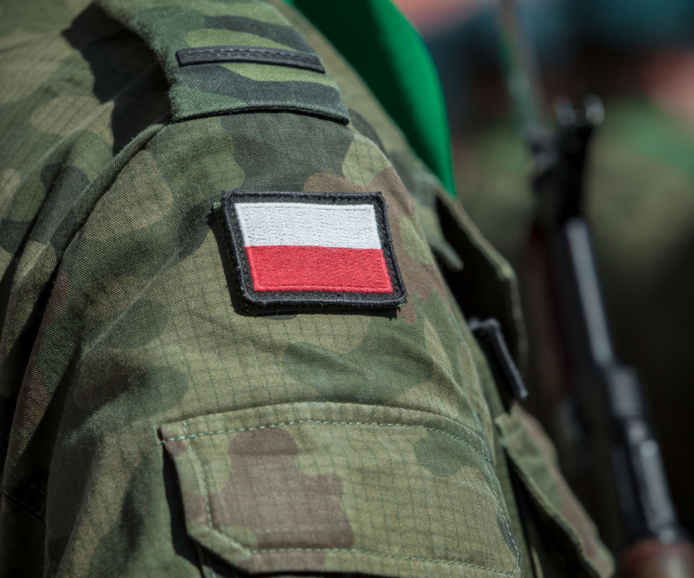 Naramiennik munduru wojskowego w kamuflażu z naszywką polskiej flagi, symbolizujący służbę i temat emerytur wojskowych, o których przeczytasz więcej na Poradnik Zdrowie. W tle niewyraźna sylwetka żołnierza z bronią.