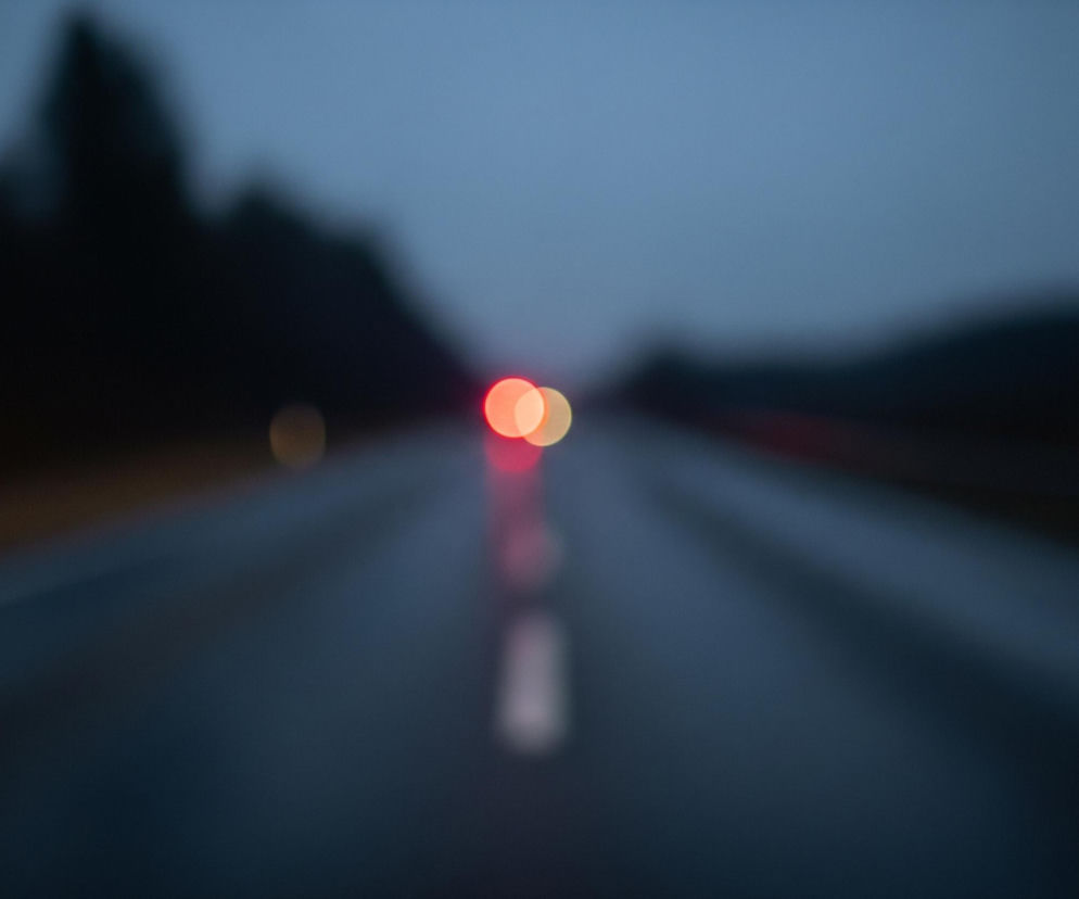 Widoczna jest rozmyta scena drogi, prawdopodobnie z perspektywy jadącego pojazdu, w ciemnym, zmierzchowym lub nocnym otoczeniu. Na środku obrazu dominuje asfaltowa jezdnia, która zdaje się uciekać w głąb kadru, z białą, rozmazaną linią podziału pasa ruchu pośrodku. W oddali, na horyzoncie, z niewyraźnej plamy światła wyłaniają się trzy wyraźne, okrągłe punkty świetlne typu bokeh: dwa czerwone i jeden jasnożółty, sugerujące światła tylne lub drogowe innych pojazdów. Po obu stronach jezdni widać zamazane, ciemne kształty, przypominające sylwetki drzew lub gęstego lasu, rozmywające się w górnej części kadru w szaro-niebieskie, pochmurne niebo.