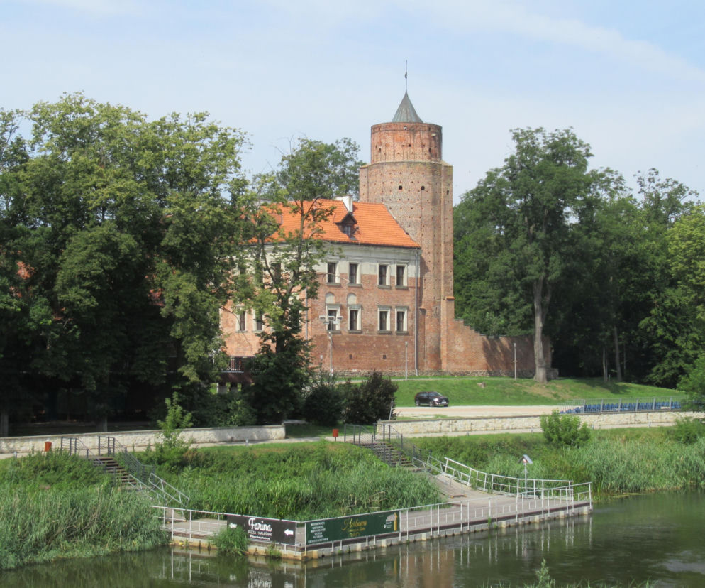 Gotycki zamek biskupów gnieźnieńskich w Uniejowie z charakterystyczną ceglaną wieżą i pomarańczowym dachem, otoczony zielonym parkiem i drzewami. Na pierwszym planie rzeka Warta z pomostem. Więcej o tym uzdrowisku przeczytasz na naszym portalu.