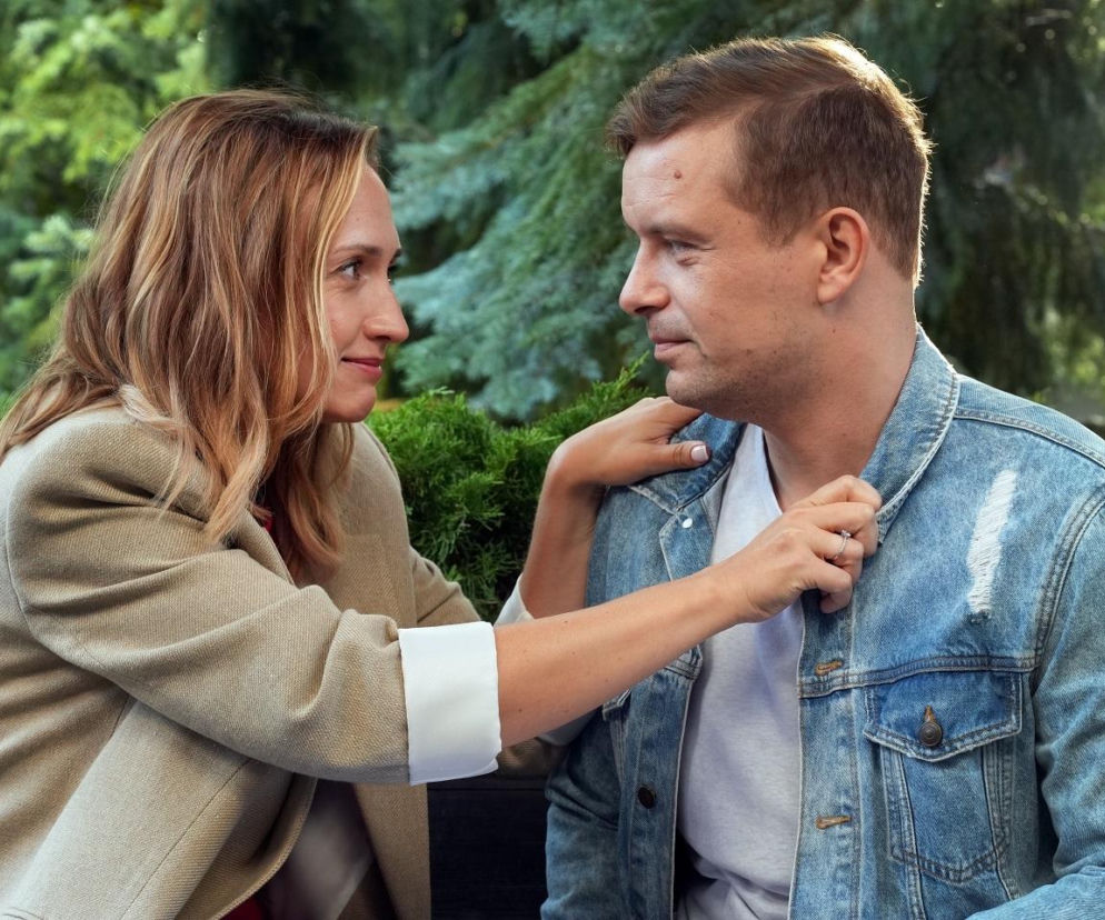 Anna Gzyra (Asia) w beżowej marynarce i Marek Molak (Hubert) w przetartej jeansowej kurtce, patrzący na siebie z czułością, w otoczeniu zielonych krzewów, na tle drzew. Ich gesty świadczą o bliskości. Więcej o ich losach w serialu Barwy szczęścia znajdziesz na Super Seriale.