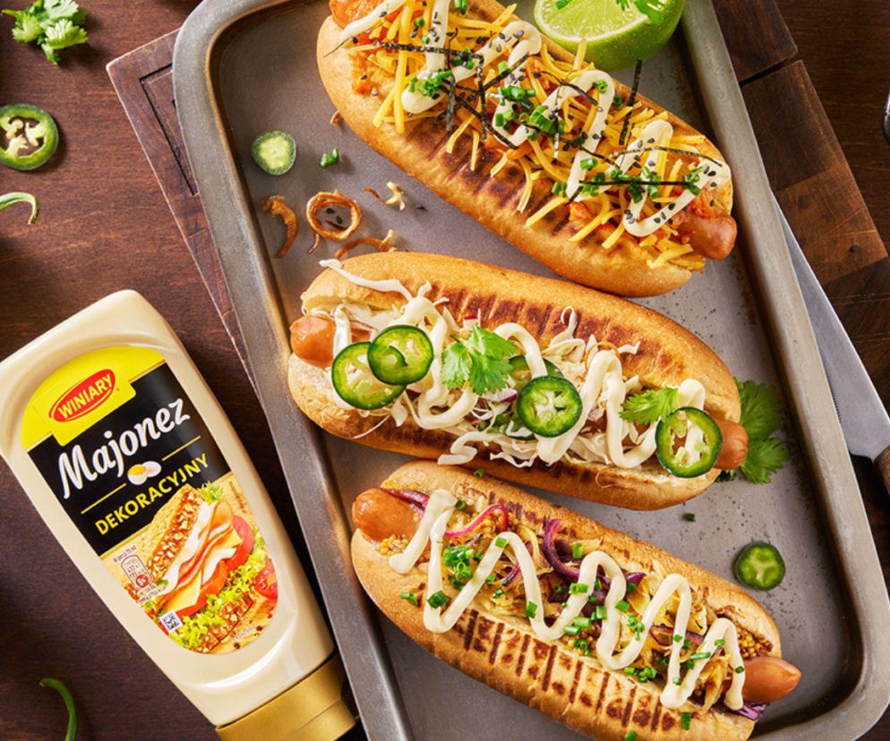 Trzy hot dogi z różnymi dodatkami i majonezem obok butelki Majonezu Dekoracyjnego WINIARY. O kampanii przeczytasz w Eska.