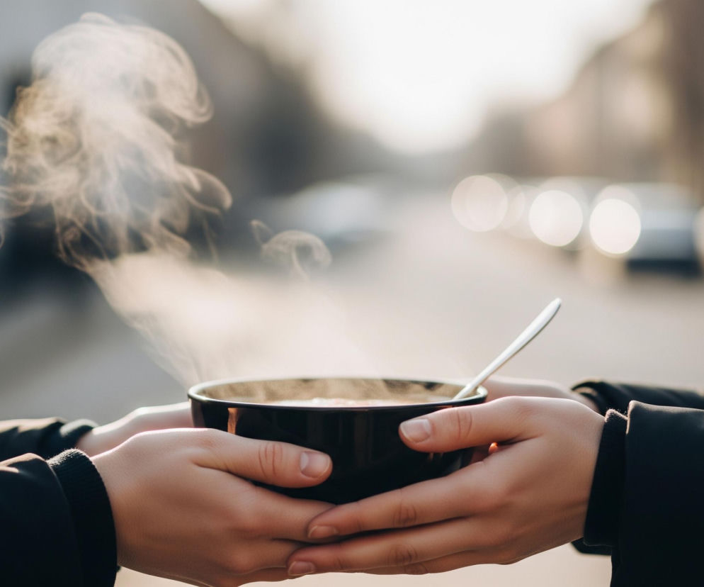 Na pierwszym planie znajdują się cztery dłonie, dwie od prawej i dwie od lewej, trzymające wspólnie ciemną miskę z gorącym napojem. Z miski unosi się biała para, a w środku widać jasnobrązowy płyn i srebrną łyżeczkę. Osoby, do których należą dłonie, mają na sobie ciemne ubrania. Tło jest rozmyte, ukazując zarysy budynków i jasne, koliste plamy świetlne, prawdopodobnie latarnie lub reflektory samochodów. Całość ma ciepłą, nieco przyćmioną kolorystykę z dominacją odcieni brązu, szarości i bieli.