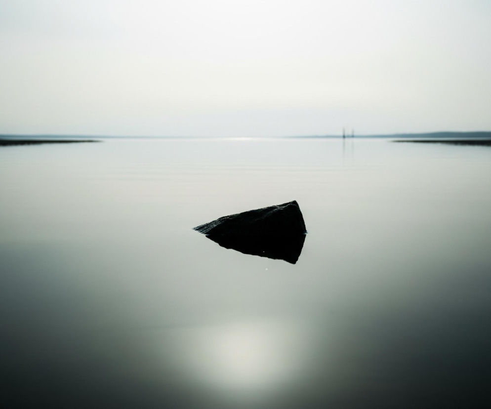 Ciemny, trójkątny kamień unosi się na spokojnej powierzchni wody, zajmując centralną część kadru. Jego górna część jest sucha, a dolna zanurzona w lekko pofalowanej tafli, której kolor przechodzi od jasnego błękitu na horyzoncie do ciemniejszego, niebiesko-zielonego w dolnej części obrazu. Na horyzoncie, rozciągającym się na całą szerokość zdjęcia, widać rozmyte, ciemne pasmo lądu lub roślinności, z jaśniejszym, rozświetlonym niebem nad nim, odbijającym się w wodzie jako podłużna, jasna poświata. Po prawej stronie horyzontu, na tle jaśniejszego nieba, widoczne są dwie niewyraźne, pionowe struktury.