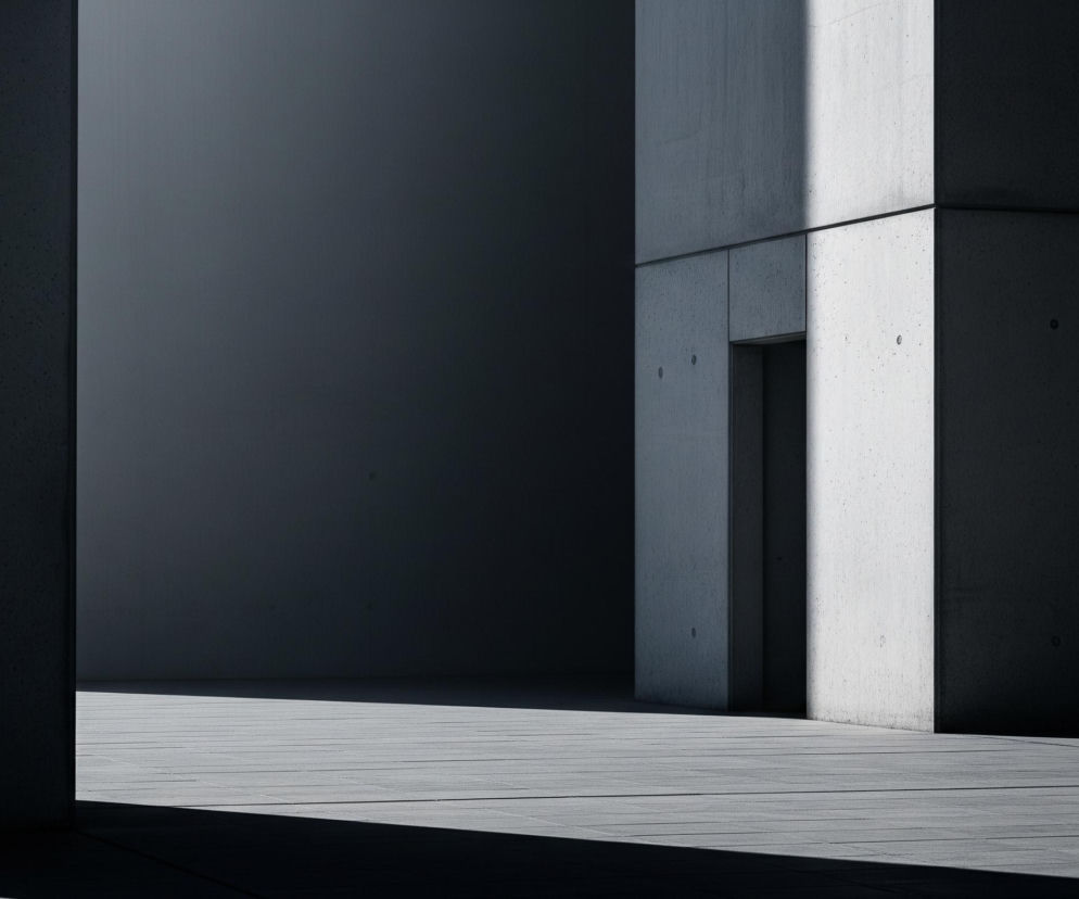 Kompozycja przedstawia geometryczne formy architektoniczne wykonane z betonu, tworząc grę światła i cienia. Po lewej stronie pionowa ściana jest pogrążona w ciemnym cieniu, a jej powierzchnia ma delikatną, nierównomierną teksturę. Prawa strona obrazu zdominowana jest przez jaśniejszą, betonową ścianę z wyraźnymi poziomymi i pionowymi fugami, która również częściowo znajduje się w cieniu, tworząc kontrastowe pasmo ciemności na swojej lewej krawędzi. Na tej ścianie widoczne są drobne okrągłe wcięcia oraz niewielkie, ciemne wgłębienie, prawdopodobnie otwór lub drzwi. Podłoże to pozioma płaszczyzna wykonana z płyt chodnikowych, częściowo oświetlona, częściowo zacieniona, co podkreśla głębię przestrzeni i minimalistyczny charakter otoczenia.