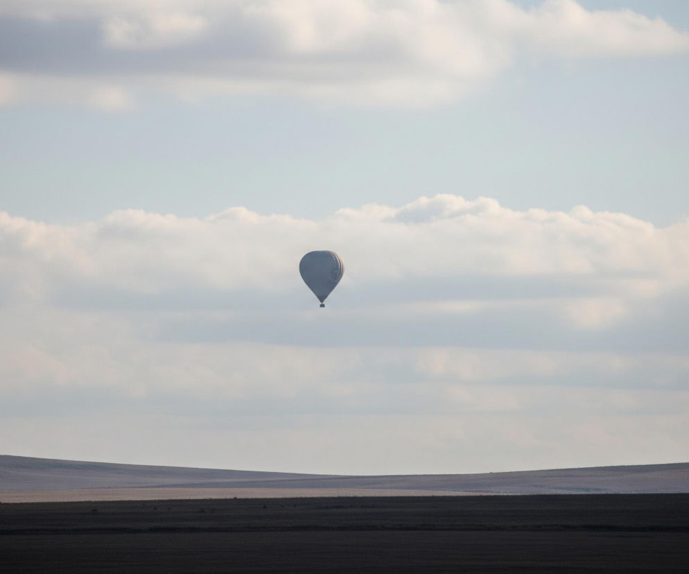 Na zdjęciu ujęty jest szary balon na ogrzane powietrze, unoszący się na tle błękitnego nieba z rozproszonymi białymi chmurami. Poniżej, w dolnej części kadru, rozciąga się horyzontalny krajobraz złożony z ciemniejszych, falujących pól, które tworzą kilka równoległych pasów w różnych odcieniach brązu i szarości. Światło rozprasza się równomiernie, tworząc lekki cień na prawej stronie balonu.