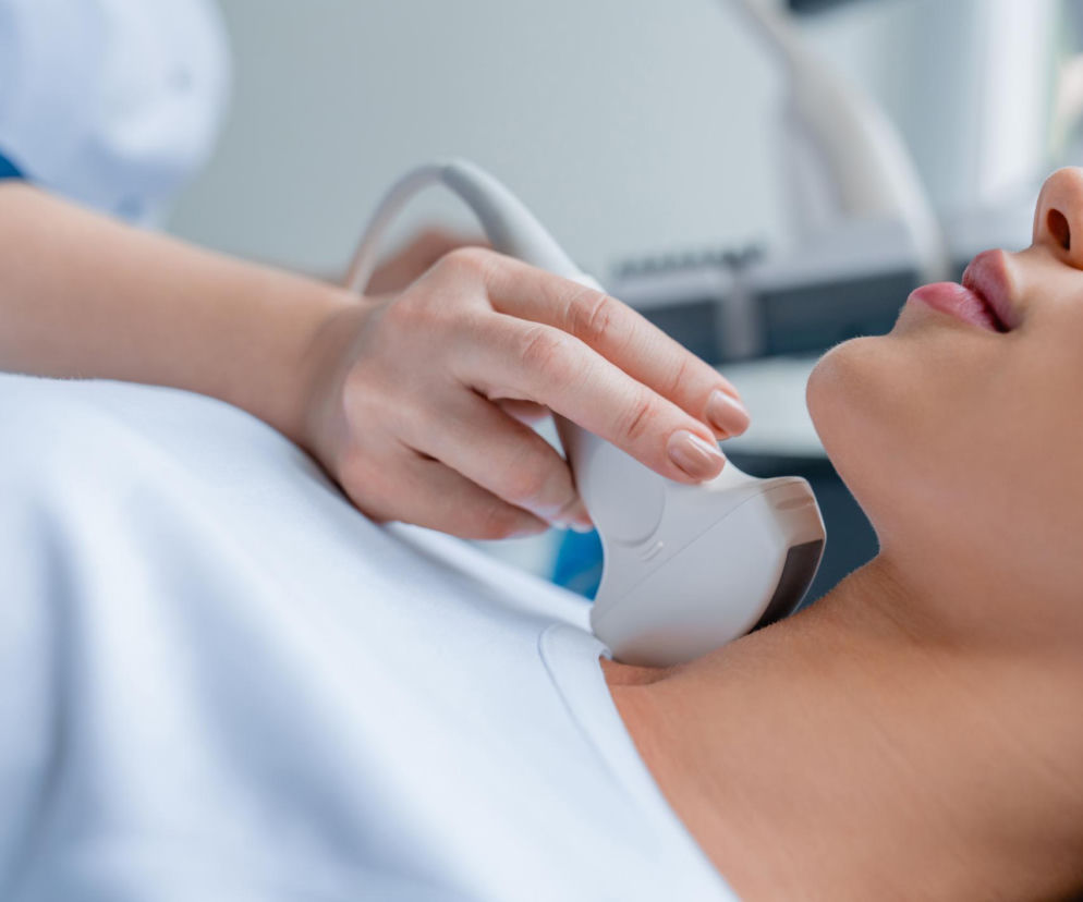 Badanie USG tarczycy u kobiety leżącej na białym prześcieradle, obrazujące diagnostykę chorób tarczycy. Lekarz wykonuje badanie za pomocą głowicy USG, analizując gruczoł szyi, o czym możesz przeczytać więcej na portalu Poradnik Zdrowie.