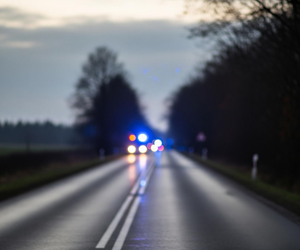 Droga asfaltowa rozciąga się w głąb obrazu, zajmując jego dolną i środkową część. Na jej powierzchni widoczne są dwie białe, ciągłe linie, które zbiegają się w oddali, tworząc perspektywę. Droga jest mokra i odbija światło, co tworzy jasne smugi. W oddali, na środku drogi, widoczne są rozmyte, intensywnie niebieskie, pomarańczowe i białe światła, sugerujące obecność pojazdów z włączonymi sygnałami świetlnymi. Po obu stronach drogi rosną drzewa i krzewy, ciemne i rozmyte, tworzące gęste pasy zieleni i brązu. Powyżej linii horyzontu rozciąga się niebo, które jest częściowo zachmurzone, z odcieniami szarości i błękitu przechodzącymi w jaśniejsze, cieplejsze barwy przy górnej krawędzi.