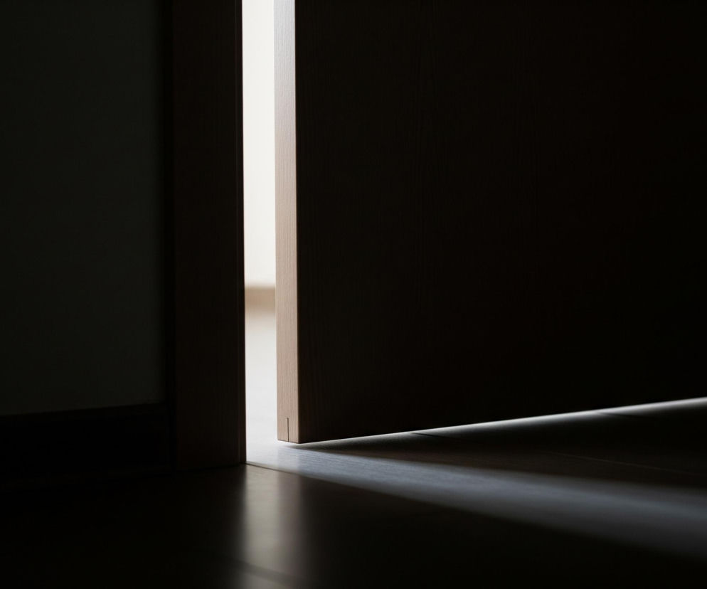 Szczelina otwartych drzwi, widoczna w pionie na środku obrazu, ukazuje jasne, pionowe pasmo światła, kontrastujące z ciemnością otoczenia. Po lewej stronie tej szczeliny znajduje się ciemna ściana, a po prawej – brązowe skrzydło drzwi, rzucające ukośny cień na podłogę. Podłoga w pomieszczeniu jest ciemna, prawdopodobnie drewniana, z jaśniejszymi fragmentami tam, gdzie pada światło zza drzwi.