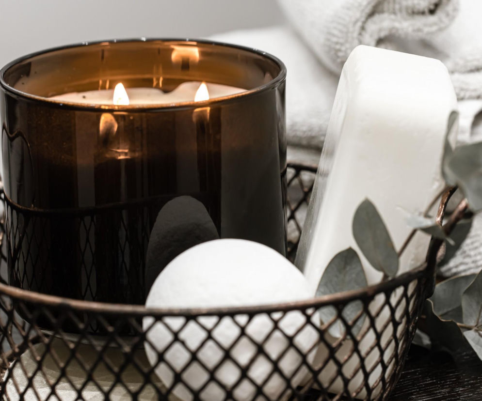 Płonąca świeca zapachowa w brązowym szkle, obok mydło i dekoracyjne liście, symbolizujące potencjalne zagrożenia dla zdrowia. Dowiedz się więcej o szkodliwości świec parafinowych i zdrowych alternatywach na Poradnik Zdrowie.