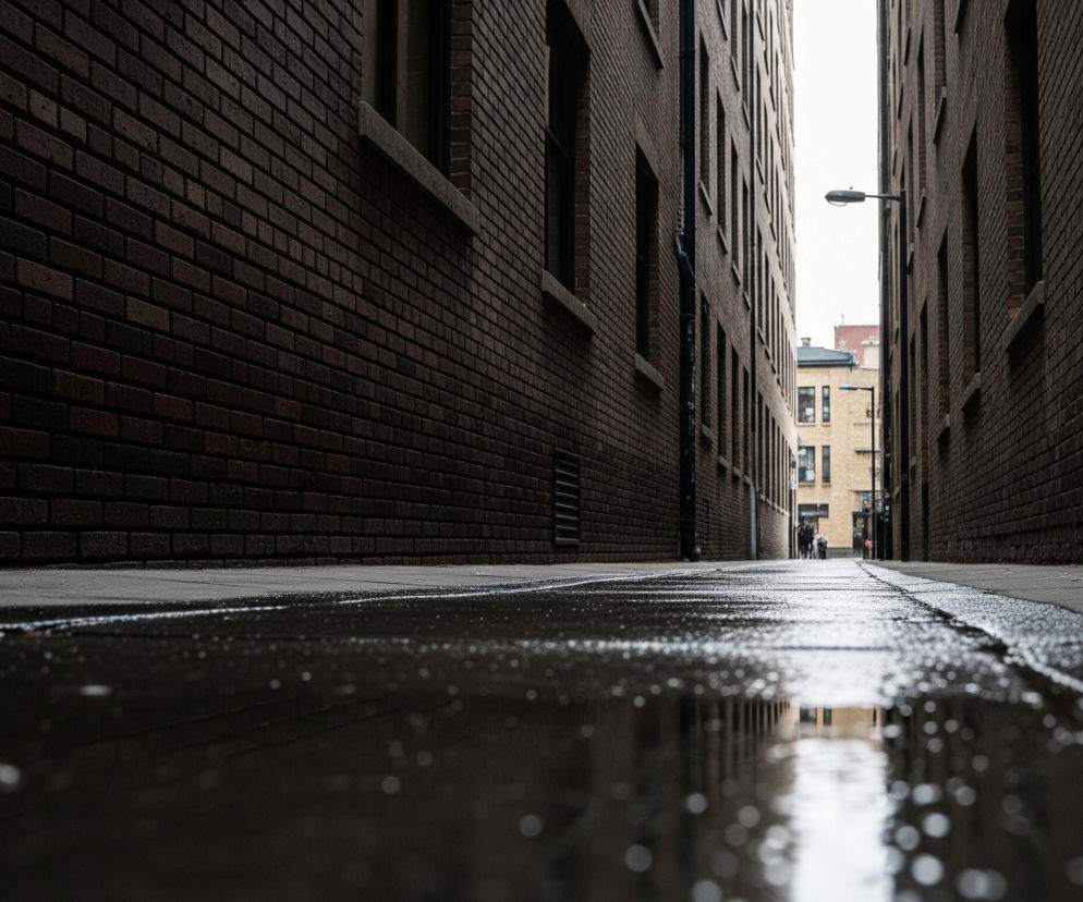 Długi, wąski zaułek rozciąga się w głąb obrazu, otoczony wysokimi, ceglanymi ścianami budynków po obu stronach. Na mokrym chodniku widać liczne kałuże i odbicia otoczenia, z jaśniejszym obszarem w oddali. W tle majaczą sylwetki ludzi oraz lampa uliczna, a niebo nad zaułkiem jest jasne.