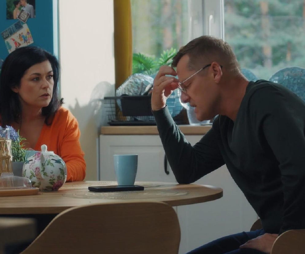 Kinga Zduńska (Katarzyna Cichopek) w pomarańczowym swetrze, z zatroskaną miną, siedzi przy stole naprzeciwko męża, Piotrka Zduńskiego (Marcin Mroczek), który w ciemnej bluzce opiera się o stół, trzymając dłoń na czole, wyraźnie przygnębiony. Zdjęcie ilustruje scenę ze złego stanu psychicznego Piotrka po spotkaniu z Pawłem, o czym można przeczytać na Super Seriale.