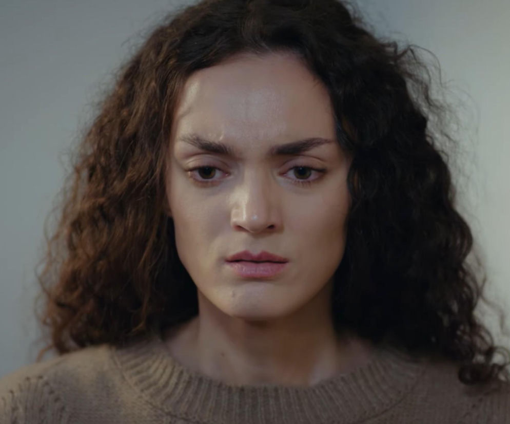 Kobieta z długimi, ciemnymi, kręconymi włosami i brązowymi oczami, spoglądająca w dół z zaniepokojeniem. Ma na sobie beżowy, dzianinowy sweter, a jej wyraz twarzy sugeruje odkrycie tajemniczego przedmiotu w serialu Dziedzictwo, o czym możesz przeczytać na Super Seriale.